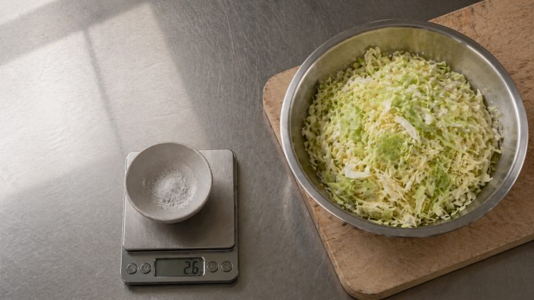 Stainless steel kitchen bench with a digital scale weighing salt in a small ceramic bowl, alongside a stainless steel mixing bowl of freshly shredded green cabbage on a wooden cutting board, neutral-warm window light from upper left.