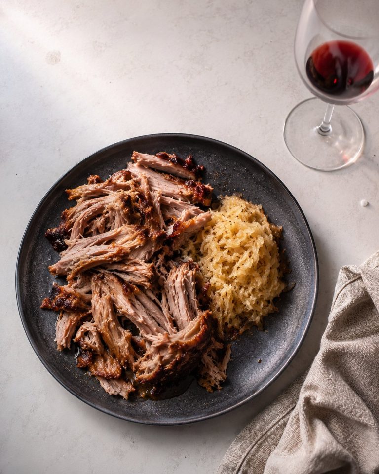 An overhead shot of a dark ceramic plate on an off-white kitchen bench. The plate holds torn slow-roasted pork shoulder with visible braised collagen, and a small spooned mound of pale gold sauerkraut. A wine glass with red wine sits partially in frame at upper right. A used linen napkin is folded at lower right. Soft window light from camera left.